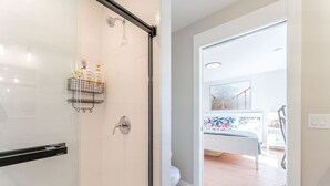 Un baño moderno con cabina de ducha de vidrio, un inodoro y una cama visible a través de una puerta abierta.