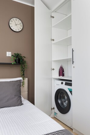 A bedroom with a bed, a wall clock, a potted plant, a washing machine, and a shelf. - Coffee Mood by Kairos Apartments (Gliwice)