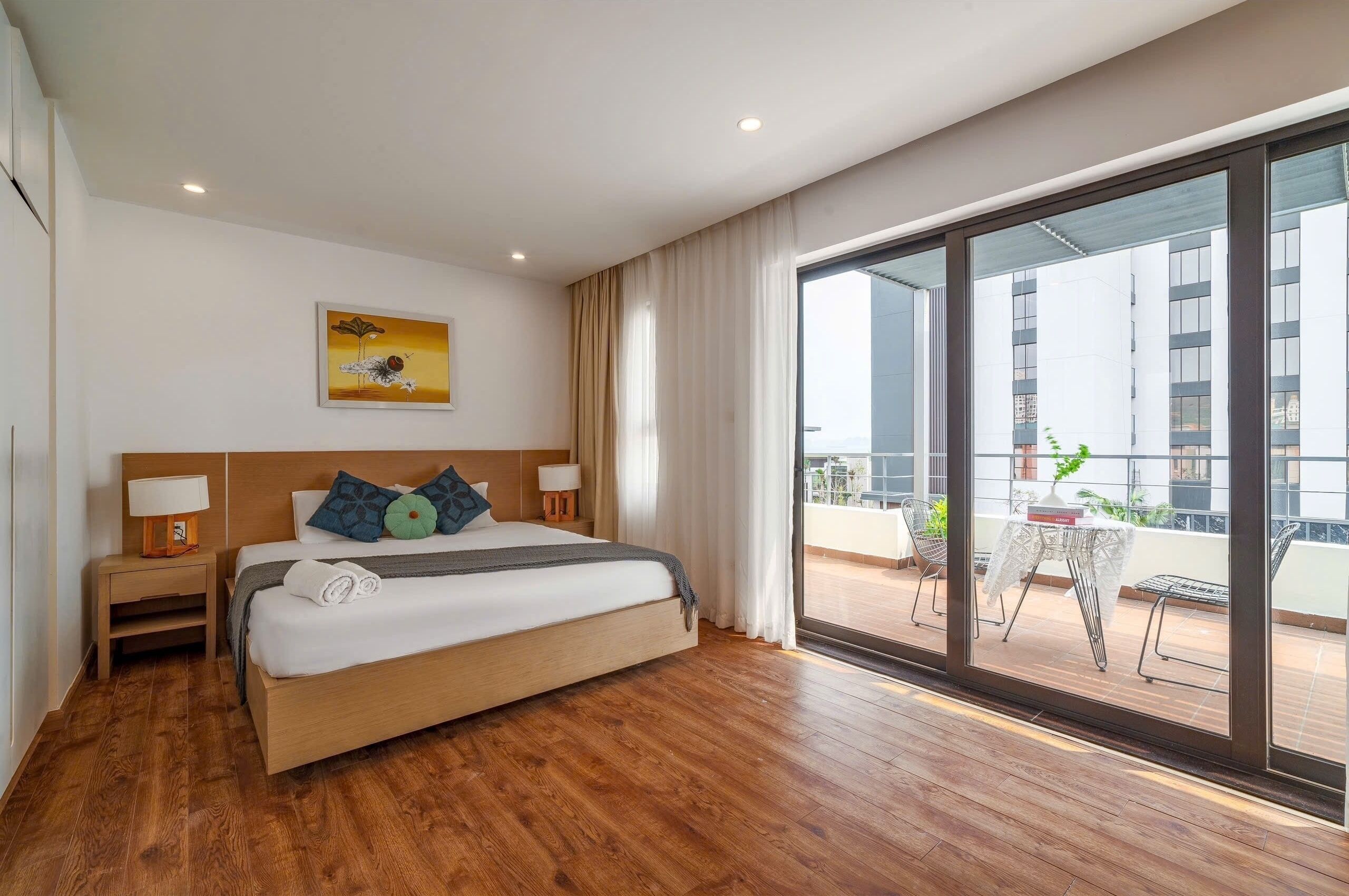 A modern hotel room with a large bed, wooden flooring, a balcony with a table and chairs, and a view of the cityscape.