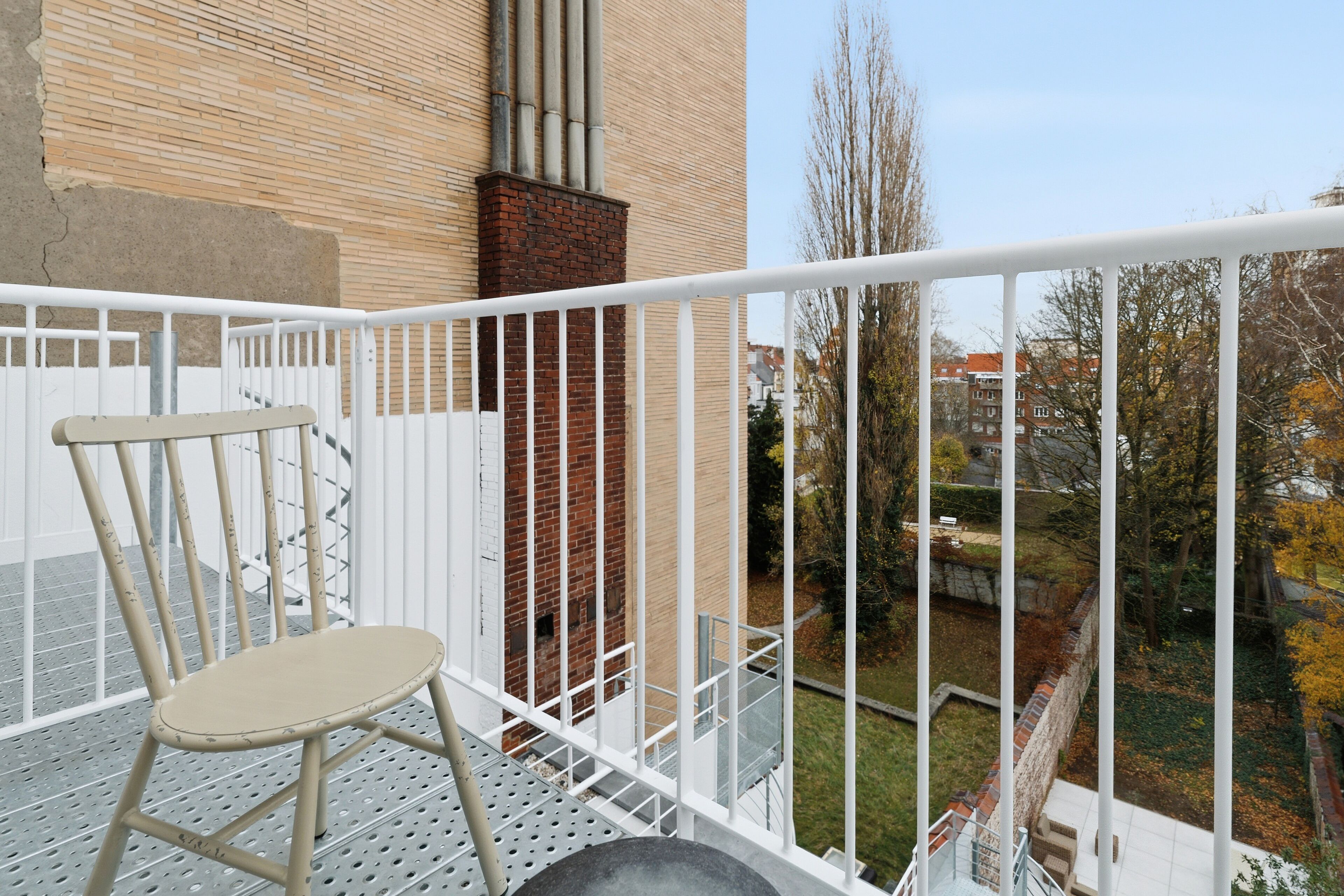 A balcony with a white metal railing, a beige chair, and a view of a brick building and trees.