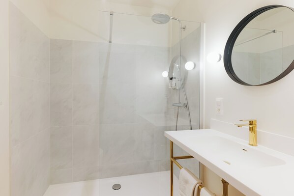 A modern bathroom with a glass-enclosed shower, a white sink with gold fixtures, and a round mirror. - Whitlock16 - Luxe Aparthotel Montgomery Square Brussels (Brussels)