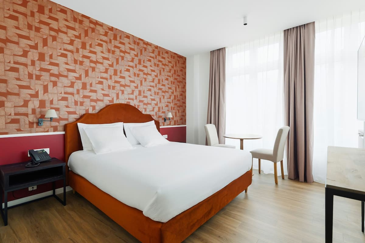 Een hotelkamer met een groot bed, een nachtkastje met een telefoon, een stoel, een tafeltje en een raam met gordijnen.