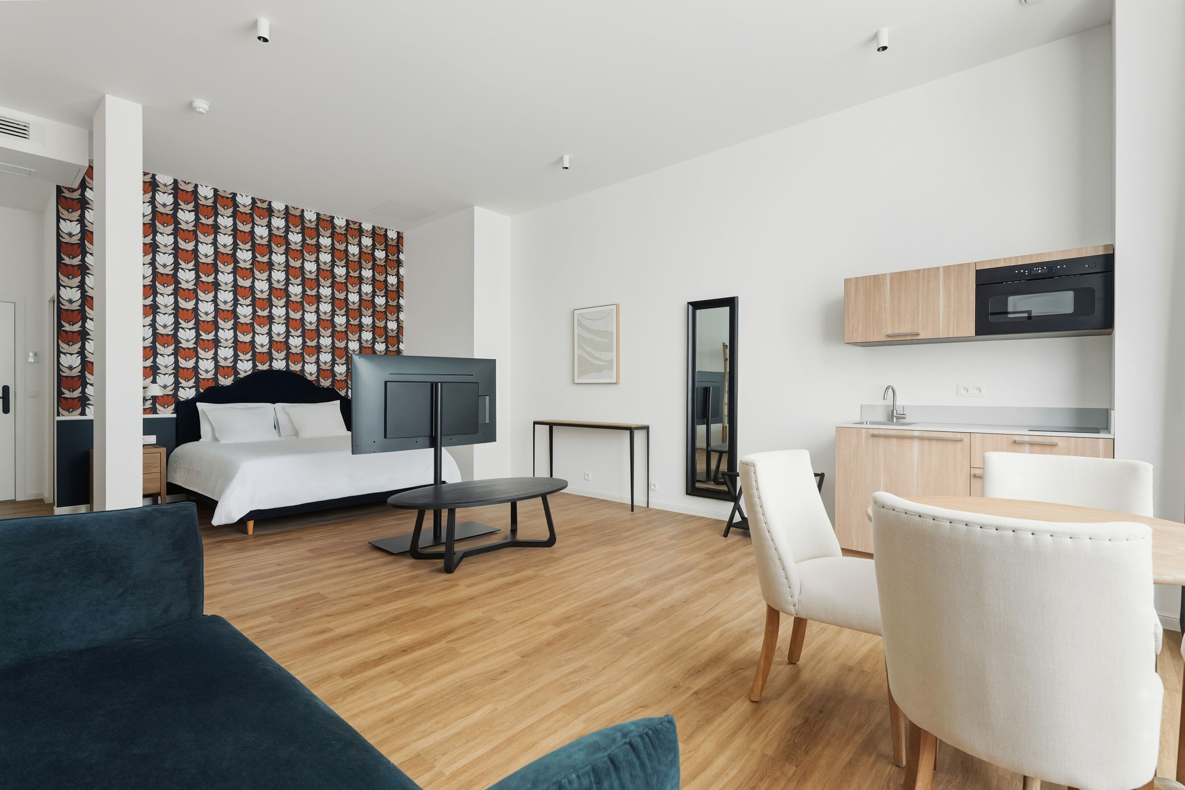 A modern hotel room with a bed, a TV, a dining table, and a kitchen area.