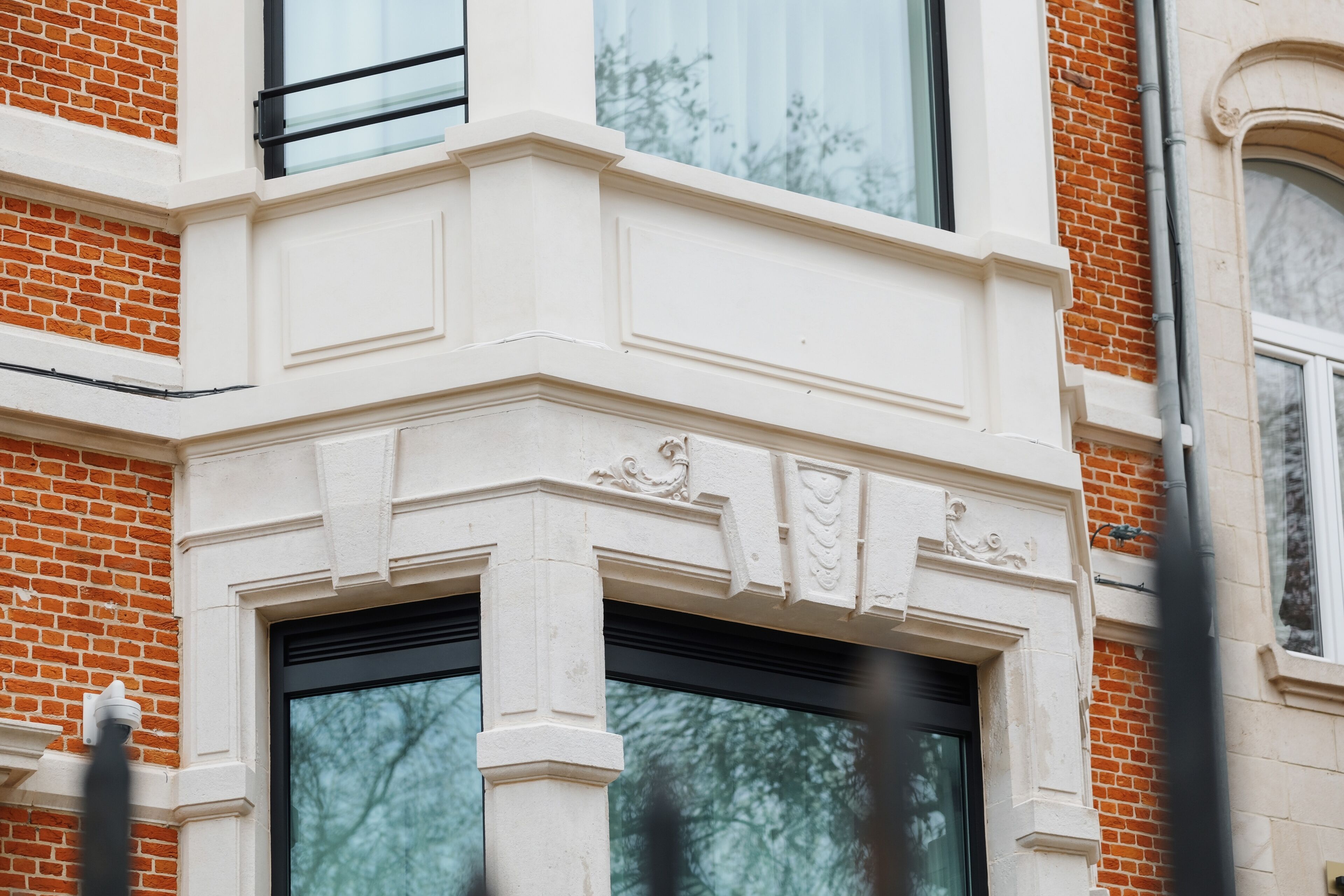 A corner of a building with a white decorative facade, a black-framed window, and a brick wall.
