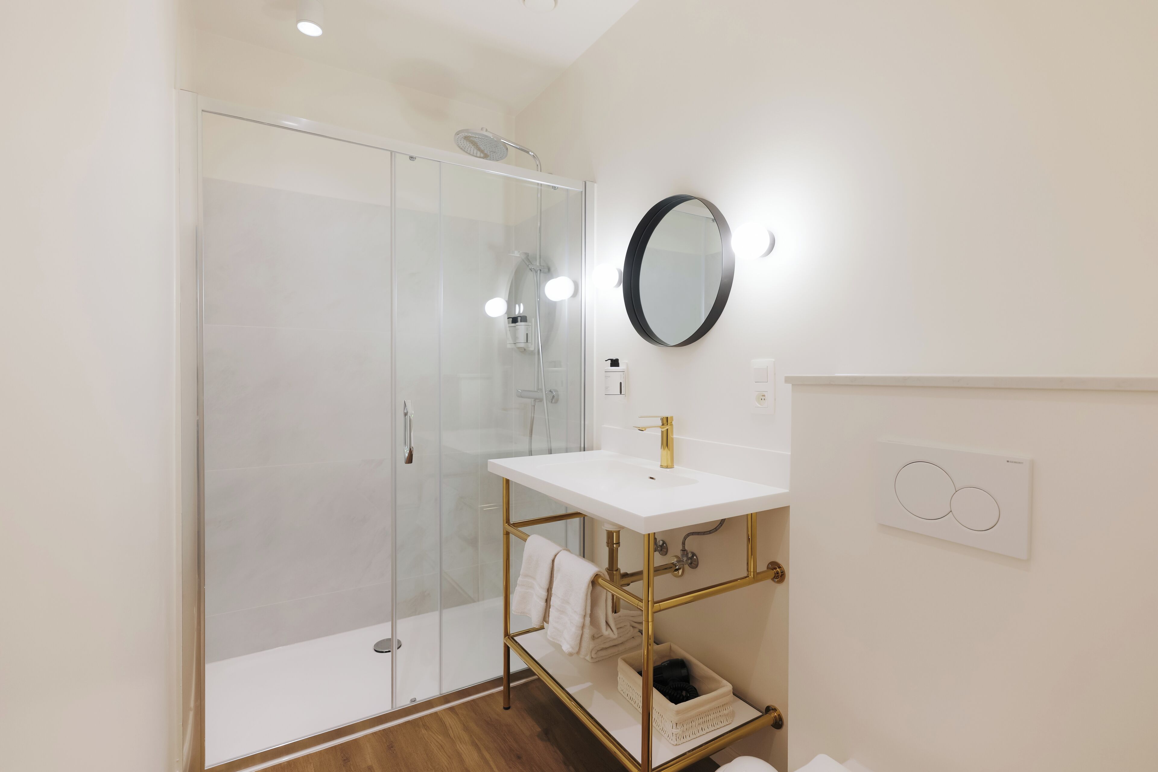 A modern bathroom with a glass shower enclosure, a white sink with gold fixtures, and a round mirror with lights.