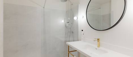 Ett modernt badrum med en duschavdelning med glasväggar, en vit tvättställ med guldfärgade detaljer och en rund spegel.