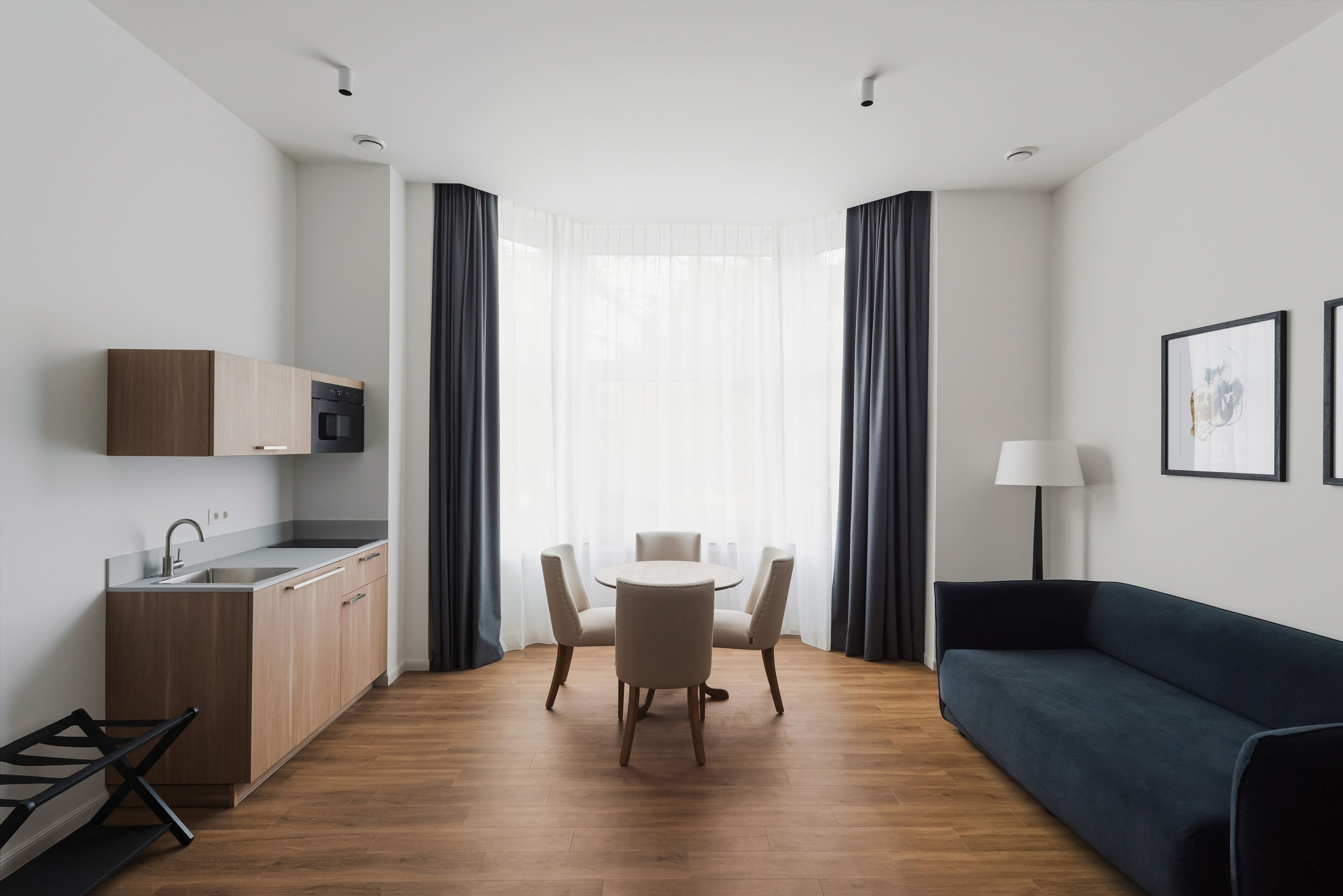 Una stanza moderna e luminosa con pavimento in legno, un tavolo da pranzo con sedie, un divano blu scuro, un angolo cottura con forno a microonde e una finestra con tende leggere.