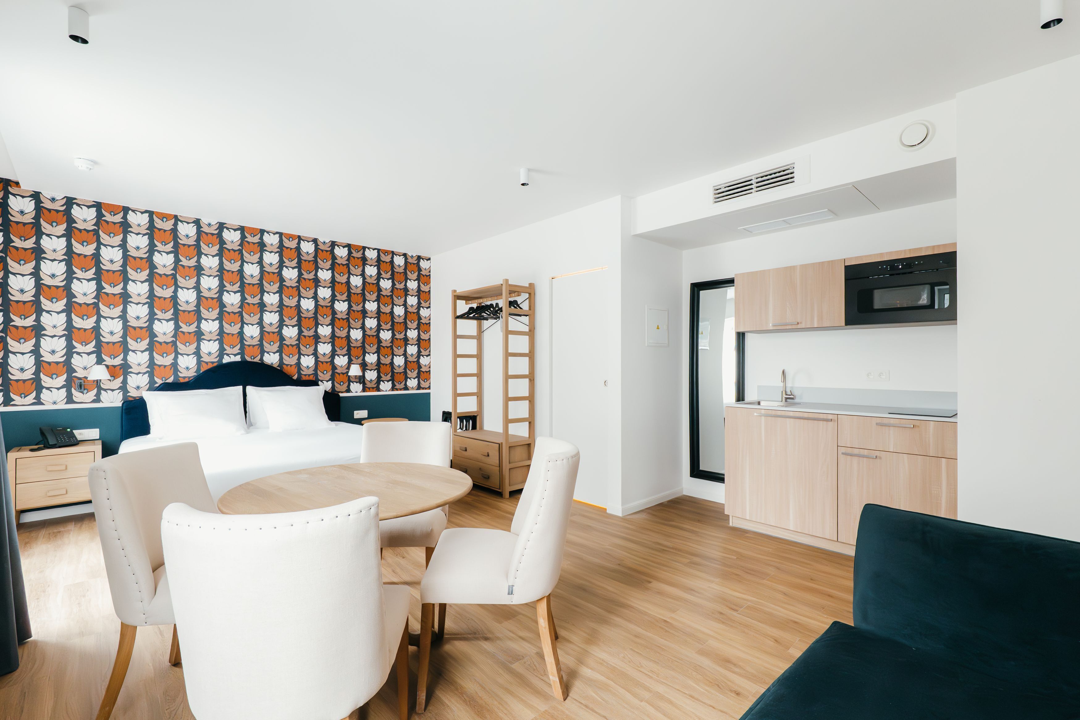 A modern hotel room with a bed, a dining table with chairs, a small kitchen area, and a wardrobe.