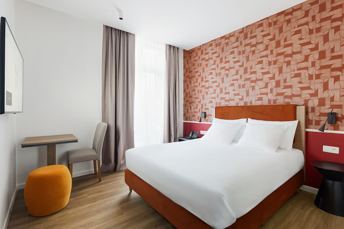 Een hotelkamer met een groot bed, een bureau, een stoel en een klein oranje poef.