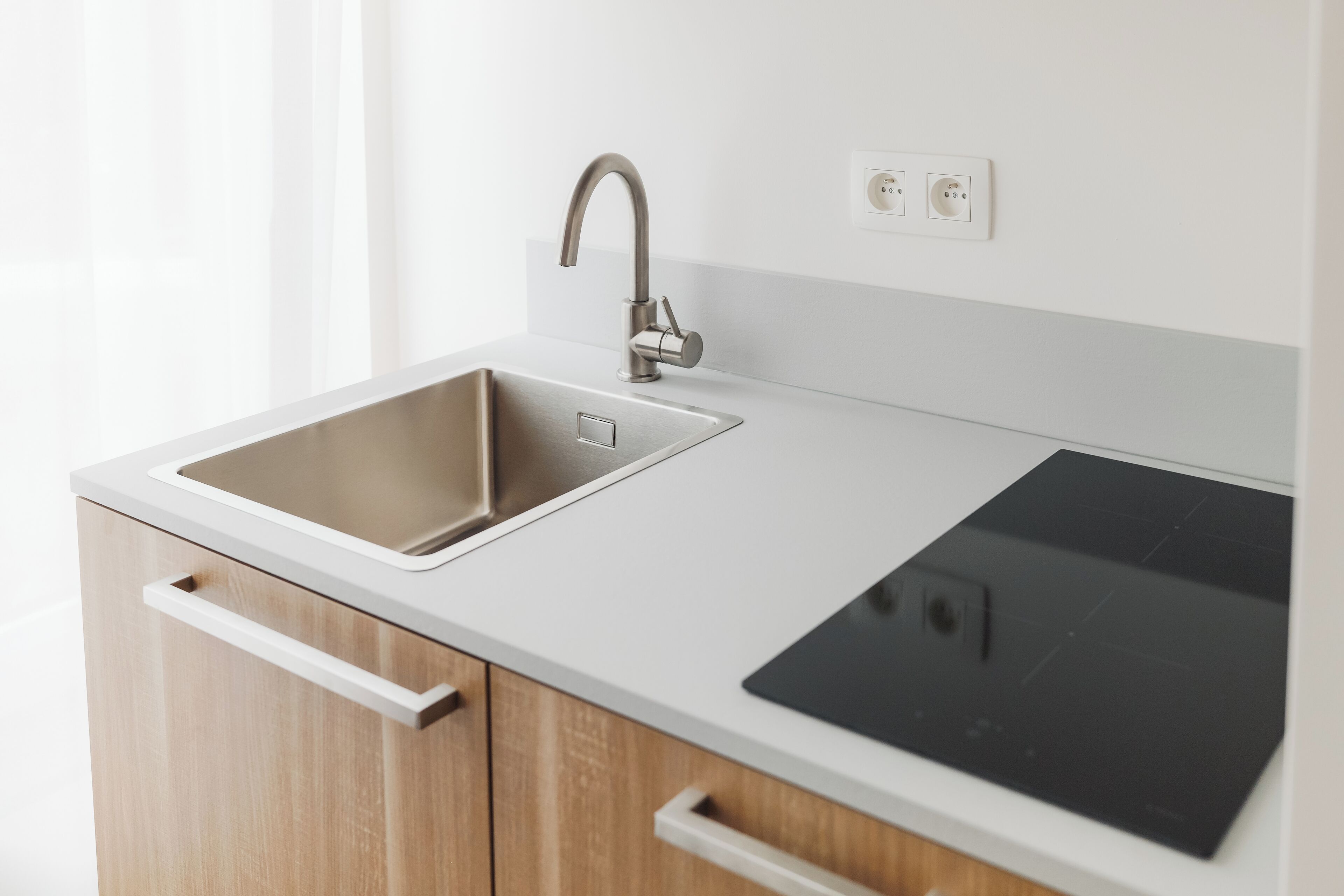 Une cuisine moderne dotée d’un évier en acier inoxydable, d’une plaque de cuisson à induction noire, de meubles en bois et d’un plan de travail blanc.