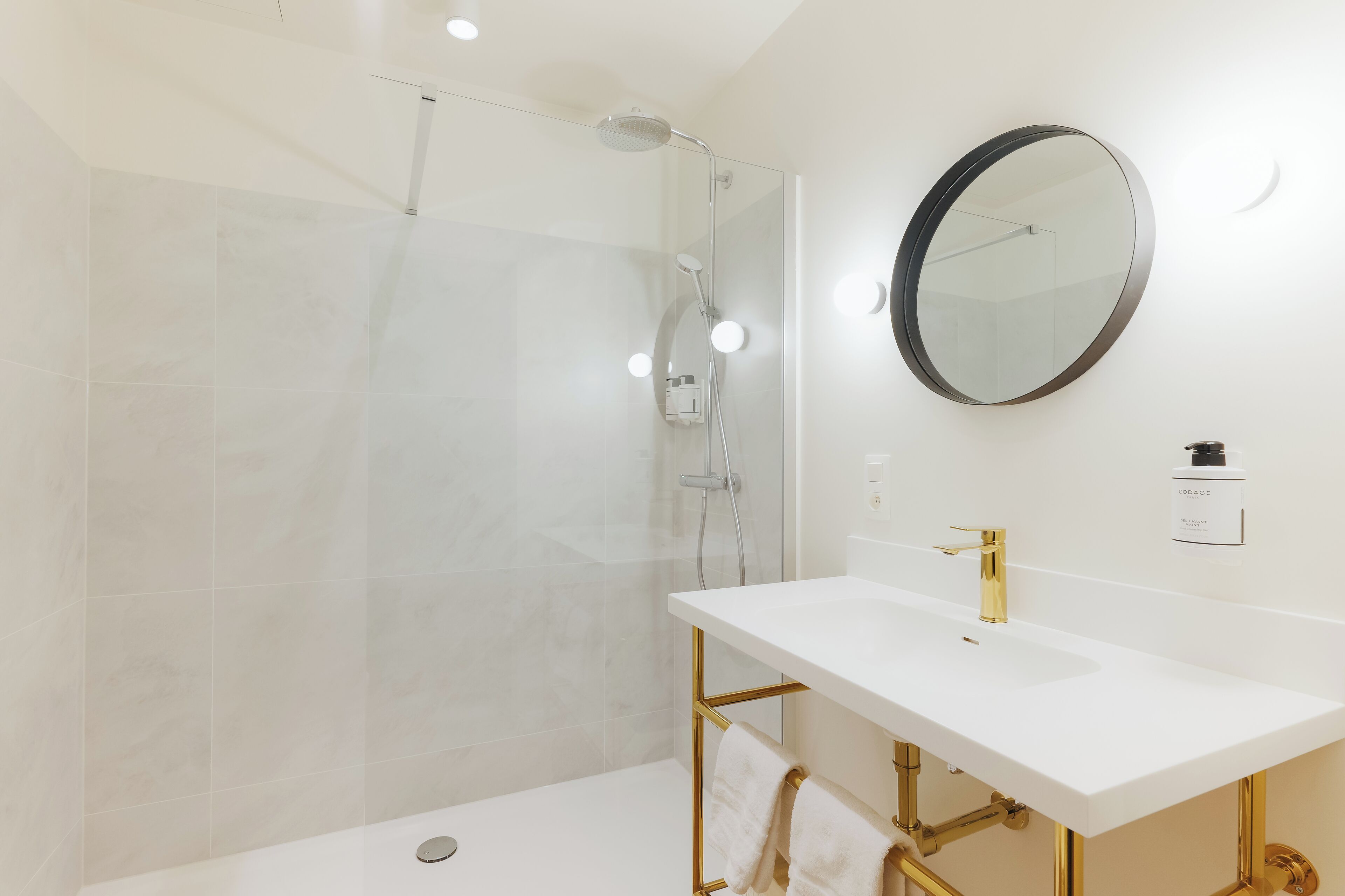 Une salle de bain moderne avec une douche à parois de verre, un lavabo blanc doté de robinetterie dorée et un miroir rond accroché au mur.