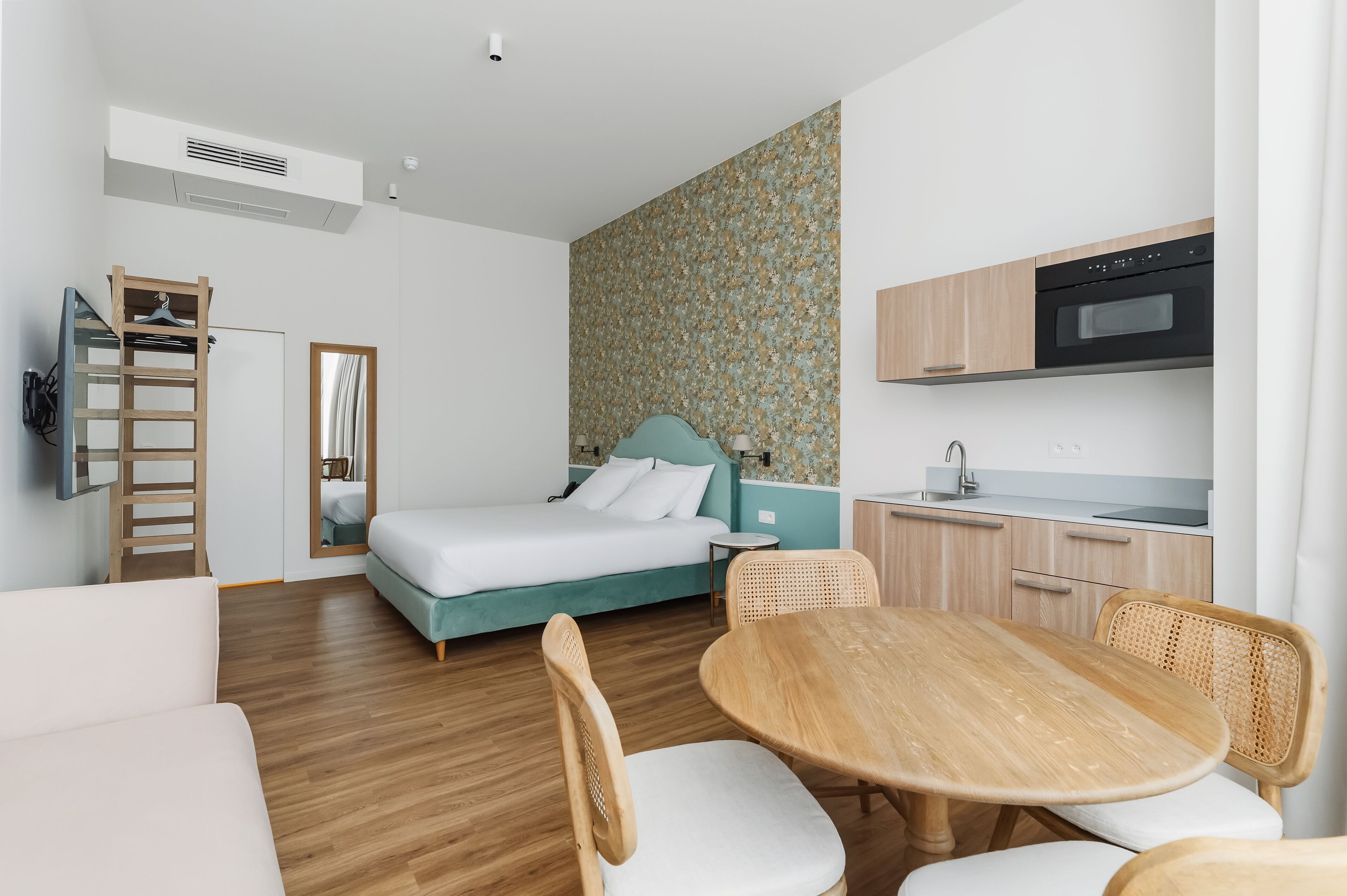 A compact room with a bed, a small kitchenette, a dining table with chairs, and a wardrobe.