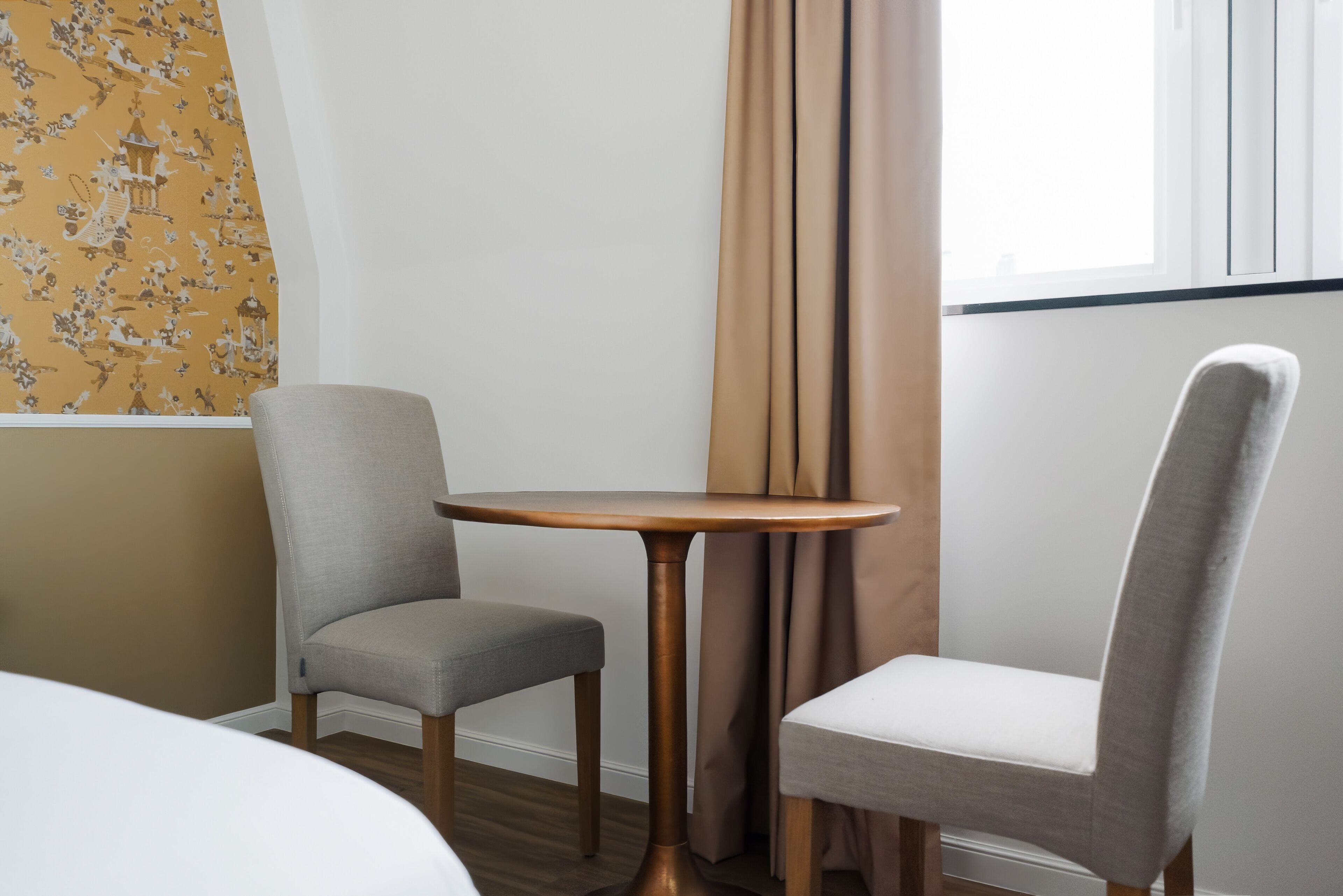 A hotel room with two grey upholstered chairs, a round wooden table, a bed, and a window with curtains.