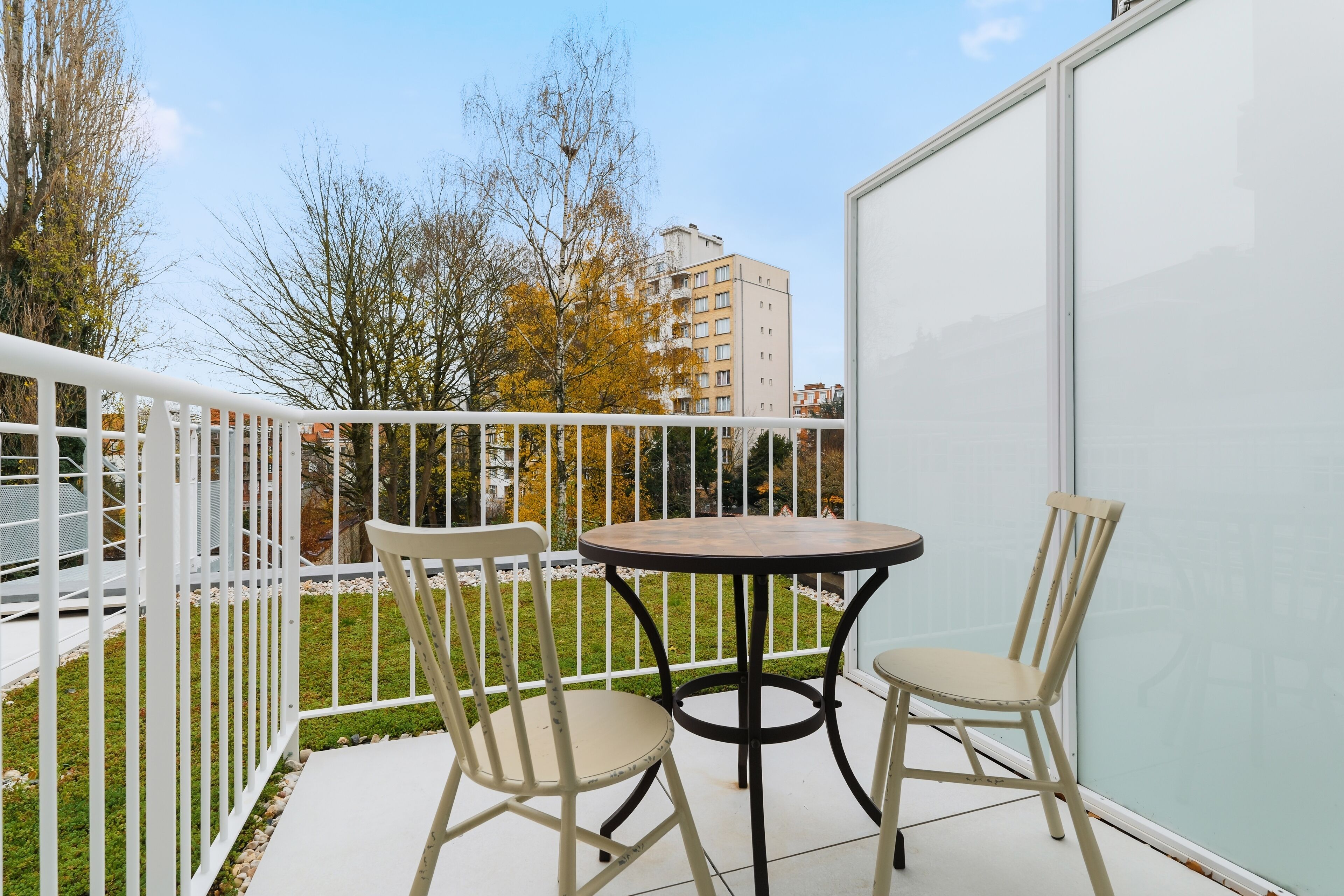 Ein Balkon mit Tisch und zwei Stühlen, einem weißen Geländer und Blick auf Bäume und Gebäude.