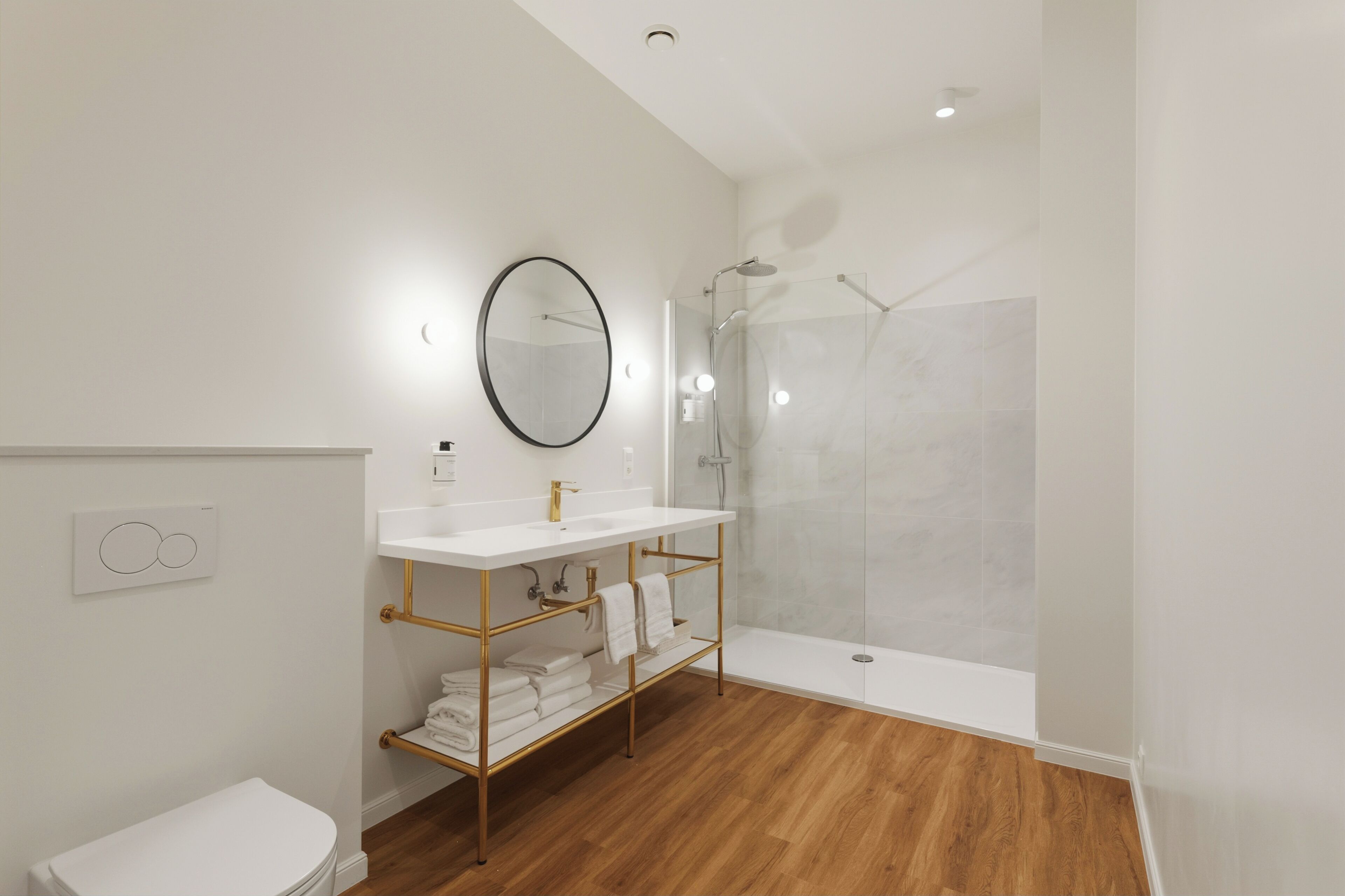 Un baño moderno con un lavabo blanco, detalles dorados, un espejo grande y una ducha con cerramiento de vidrio.