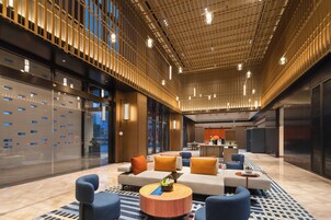 Un vestíbulo espacioso con diseño moderno, que presenta una zona de estar central con sofás y sillones, un suelo estampado y un techo con luminarias lineales.