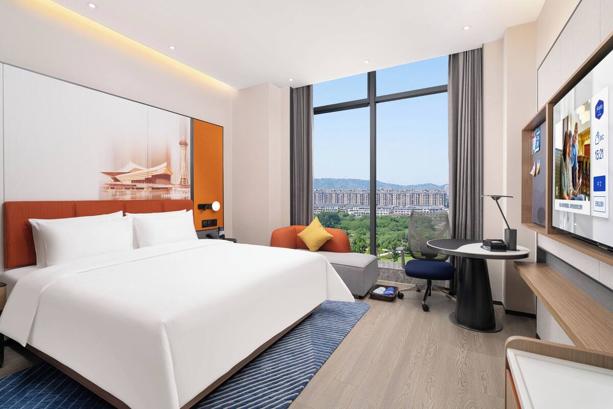 A modern hotel room with a large bed, a desk, and a view of a cityscape.