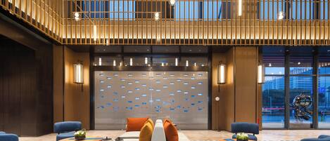 A modern hotel lobby with a seating area, a reception desk, and a large window with a view of the cityscape.