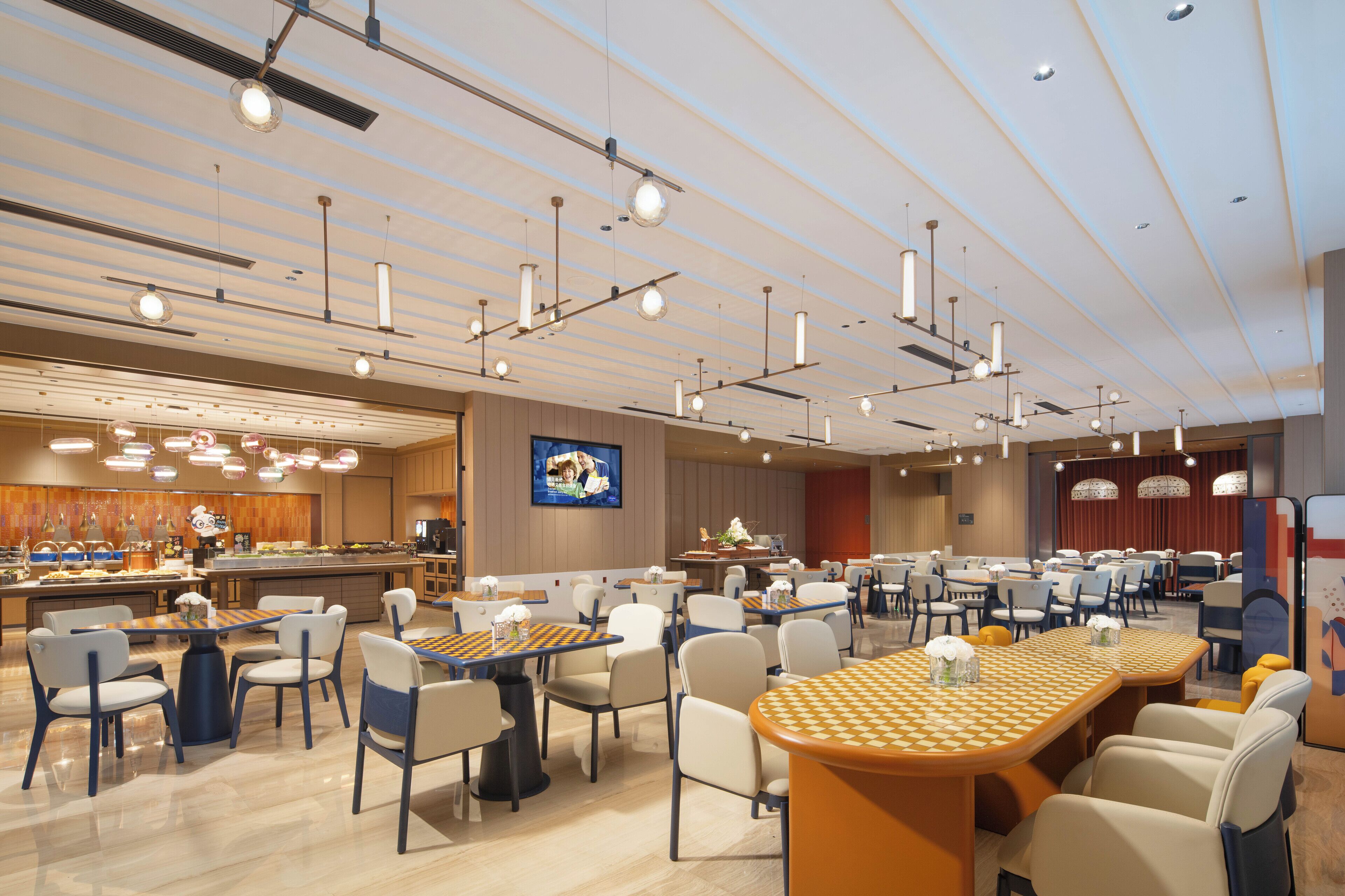 A spacious hotel lobby with multiple dining areas, a central bar, and a large screen displaying a program.