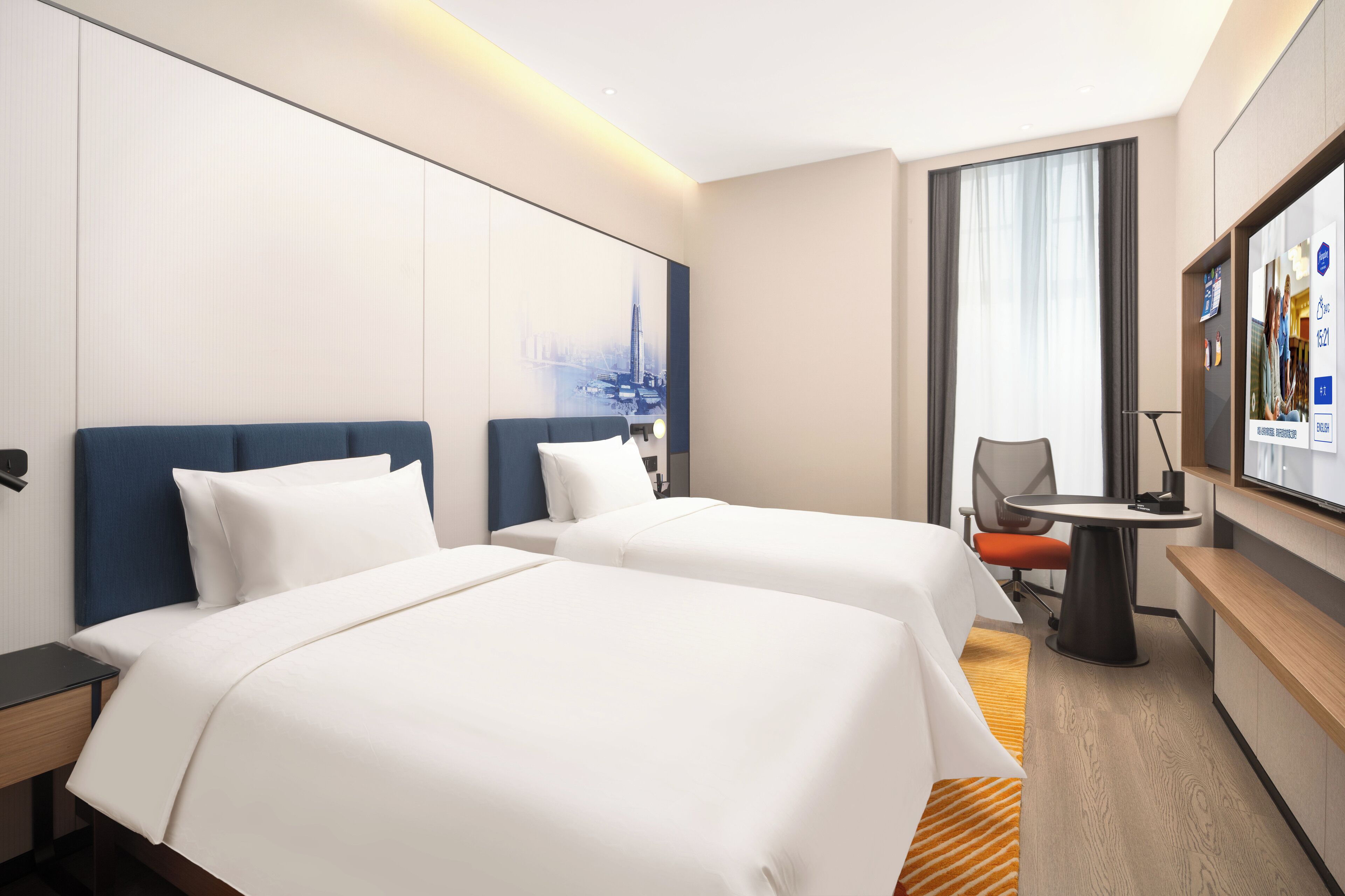Quarto de hotel com duas camas, uma escrivaninha, uma cadeira, televisão e uma janela grande com cortinas.