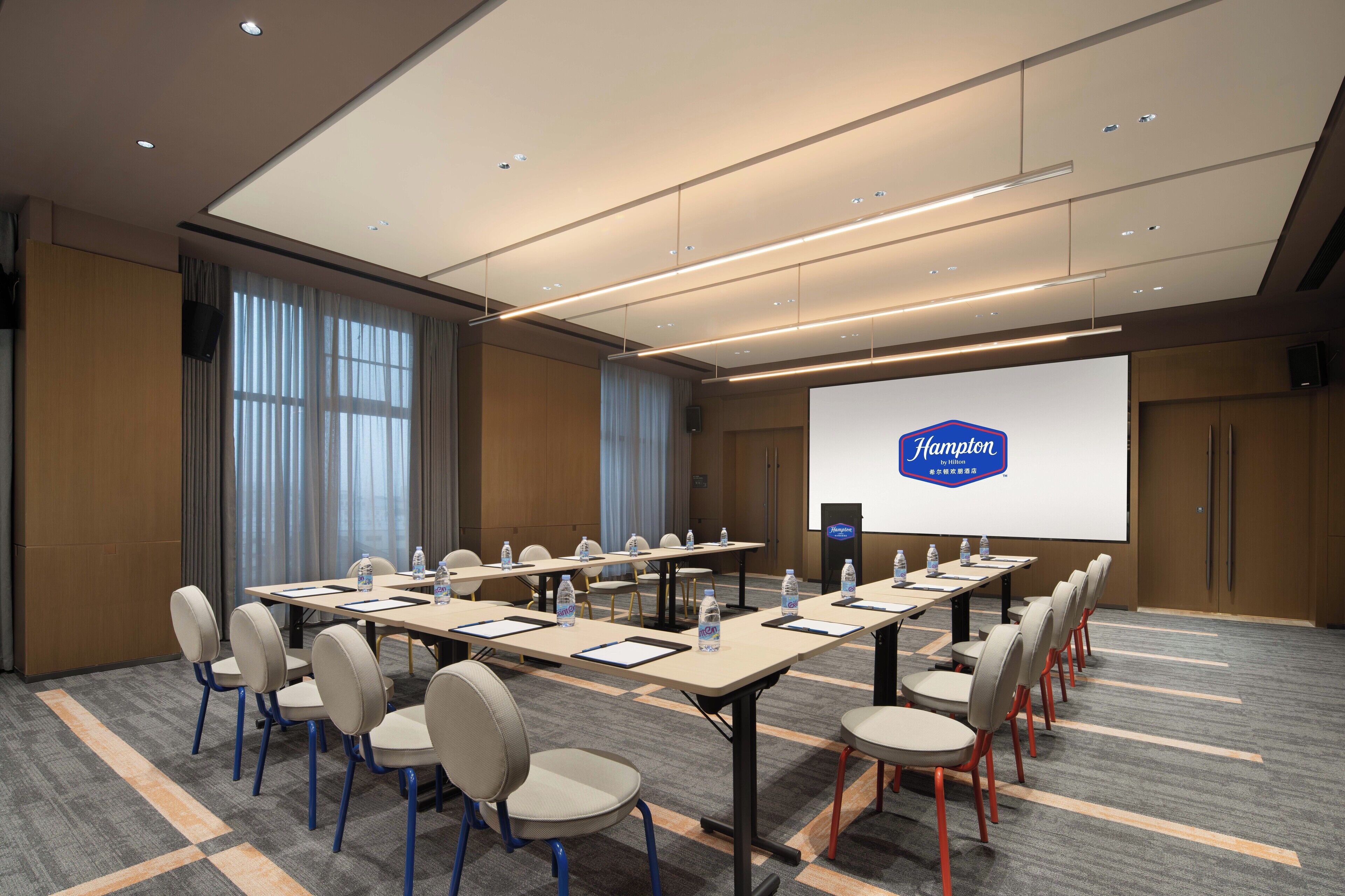 Un salón de conferencias con una pantalla grande mostrando el logotipo de Hampton, varias mesas con sillas y botellas de agua sobre las mesas.