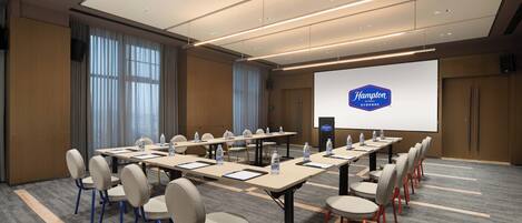 Un salón de conferencias con una pantalla grande mostrando el logotipo de Hampton, varias mesas con sillas y botellas de agua sobre las mesas.