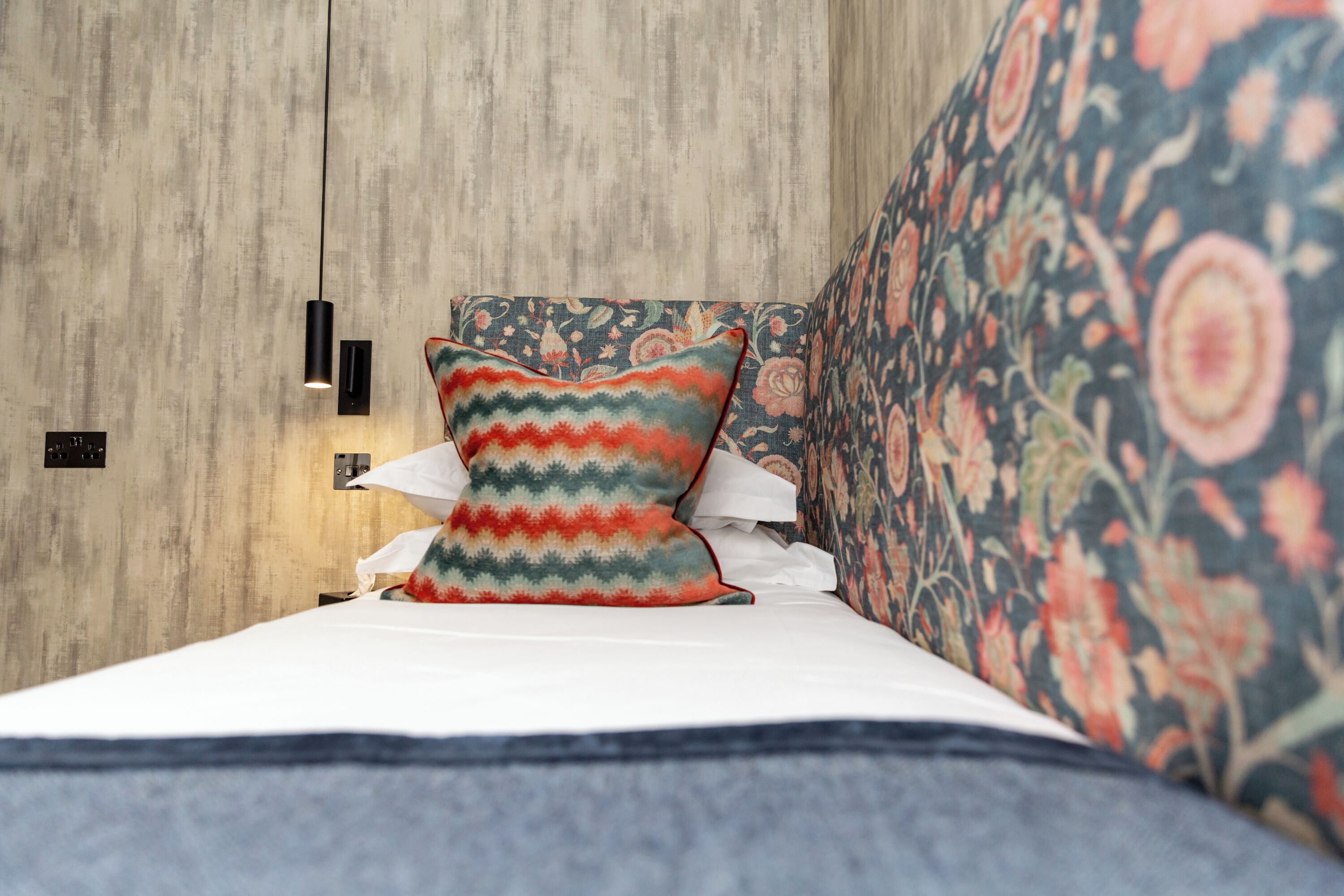 A bed with a patterned pillow, a floral upholstered headboard, and a wall-mounted light fixture.