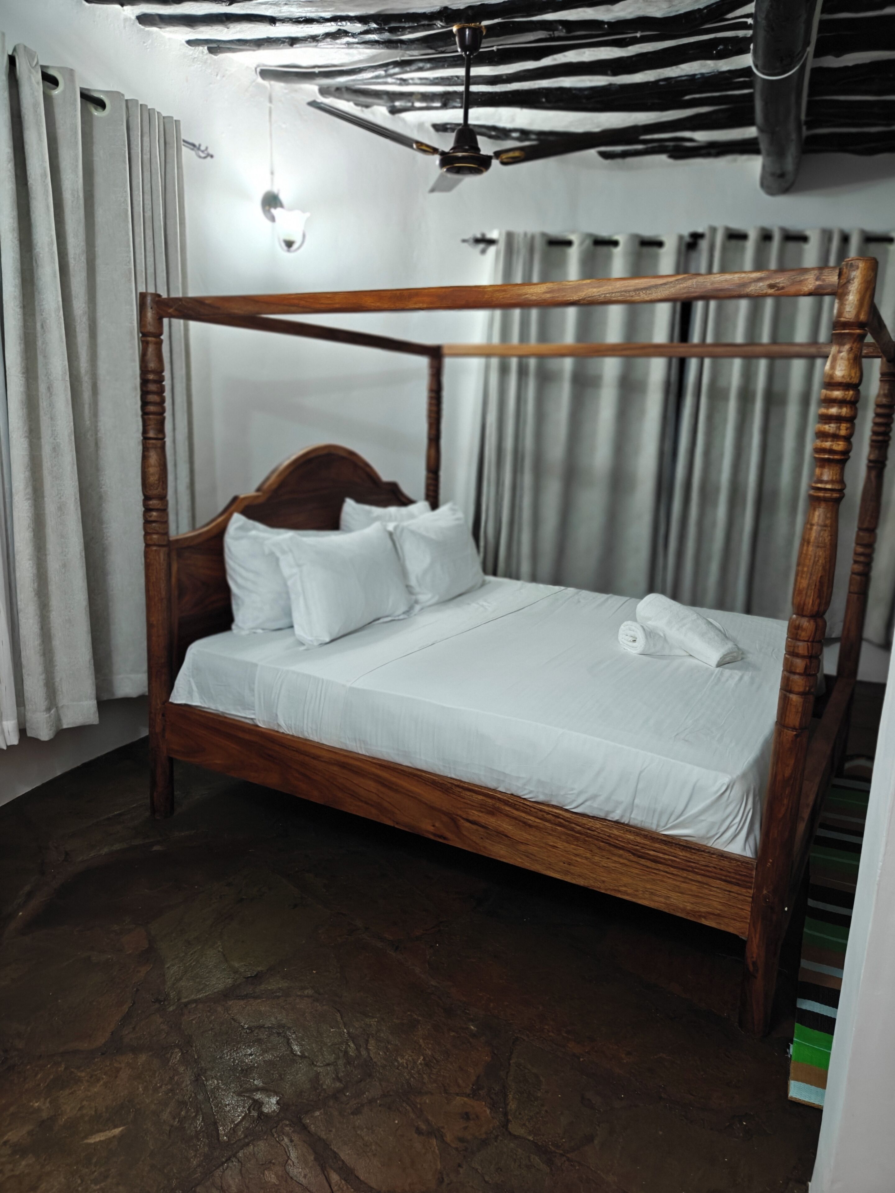 A four-poster bed with white linens, a wooden headboard, and a stone floor.