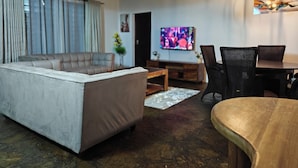 Een moderne woonkamer met een bank, eettafel, stoelen, een televisie en een plafond met zichtbare balken.