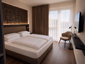 A hotel room with a large bed, a desk, a chair, a flat-screen TV, and a window with curtains. - In2 The Hotel  (Xanten)