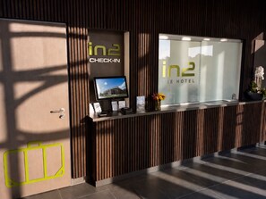 A reception desk with a monitor, a glass window displaying the hotel name, and a plant. - In2 The Hotel (Xanten)