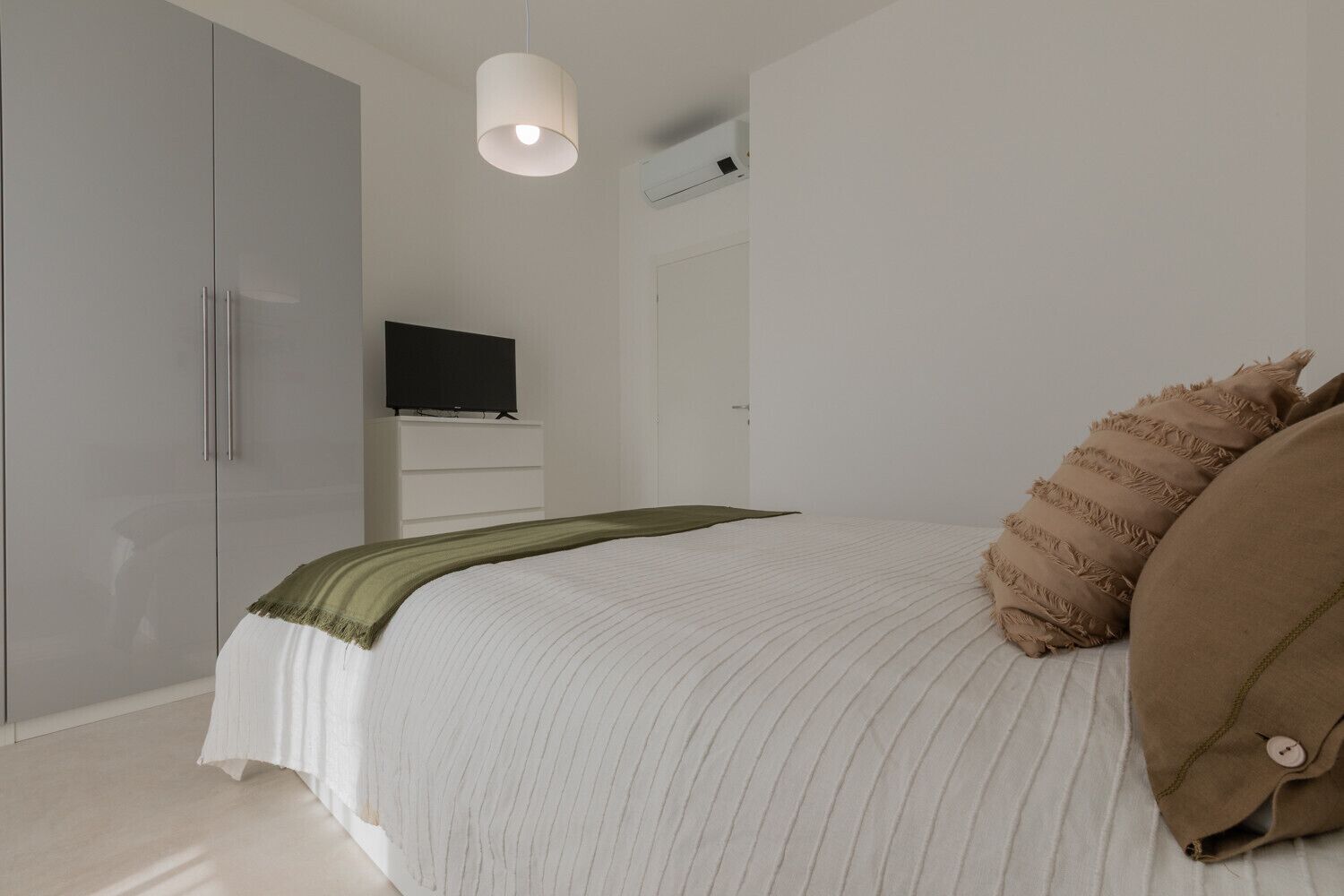 A bedroom with a bed, a television on a stand, a wardrobe, and a hanging light fixture.
