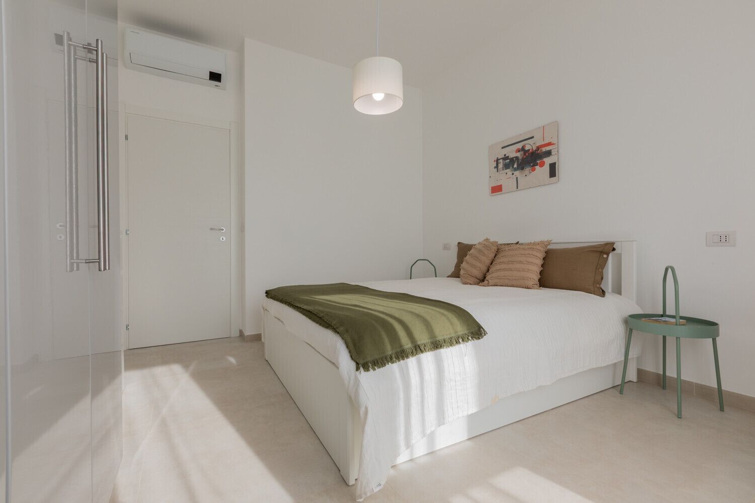 Een minimalistische slaapkamer met een bed, een nachtkastje, een groene fauteuil en een kunstwerk aan de muur.