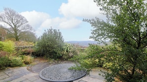 A round metal table with a decorative base, surrounded by stone paving, set in a garden with various plants and trees. - The Farmhouse (High Peak)