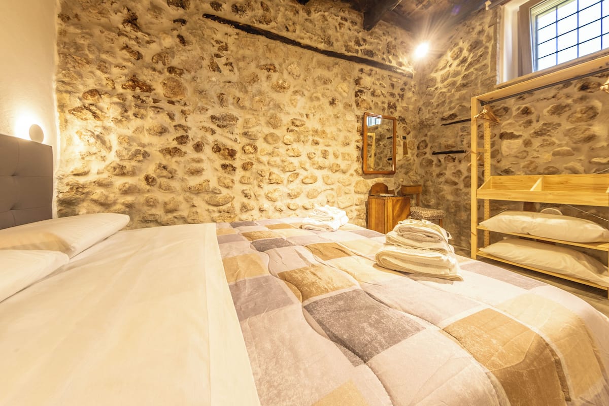 A room with a stone wall, a bed with a quilt, a wooden shelf with pillows, and a mirror on the wall.