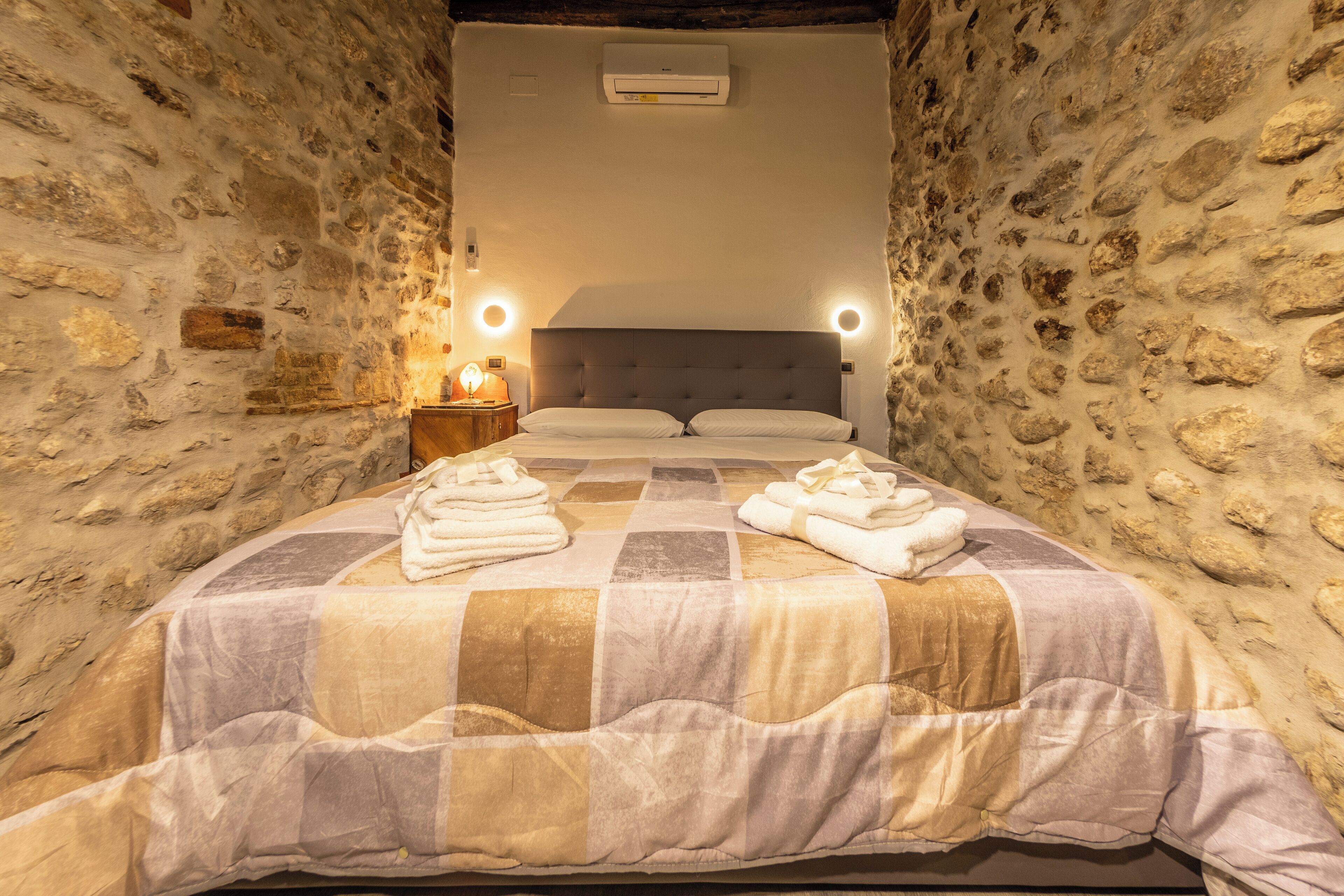 A stone-walled bedroom with a bed, two bedside lamps, and a wall-mounted air conditioner.