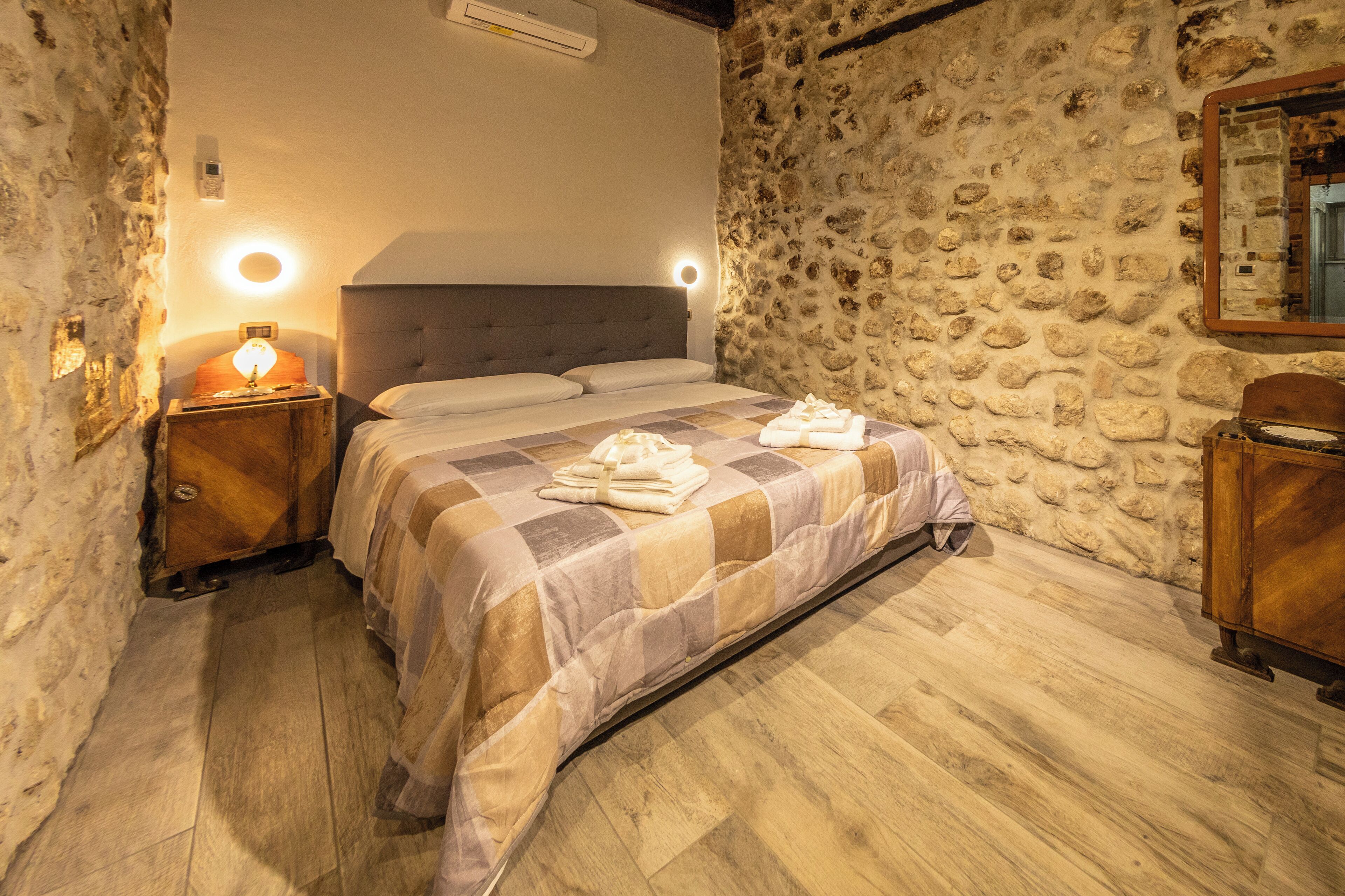 A bedroom with a stone wall, a bed with a checkered bedspread, a bedside lamp, and a wooden nightstand.