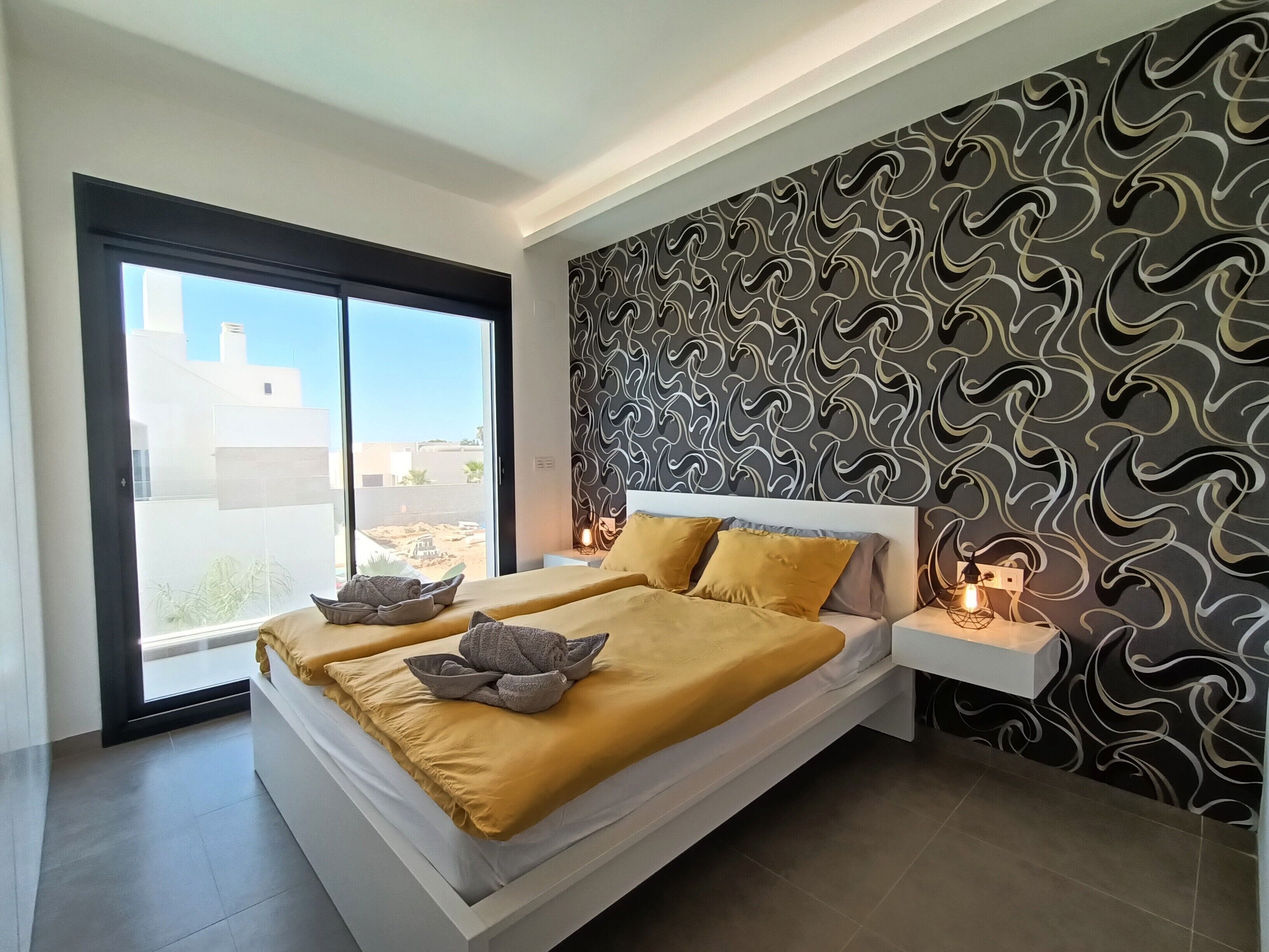 A modern bedroom with a large bed, a sliding glass door, a patterned wall, and bedside lamps.
