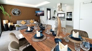 Een moderne eetruimte met een houten tafel voor zes personen, een bank met kussens, een wandgemonteerde televisie en een keuken op de achtergrond.