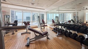 A gym with exercise equipment, large windows, and a view of buildings. - StayShort - Bright 1BR in JLT with Stunning Lake Views! (Dubai)