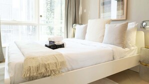 A modern hotel room with a large bed, a bedside table, a lamp, and a view of the city through large windows. - StayShort - Bright 1BR in JLT with Stunning Lake Views! (Dubai)