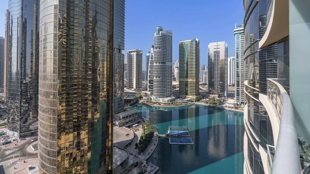 A high-rise building with a curved glass facade, a waterfront with a boat dock, and other modern skyscrapers in the background. - StayShort - Bright 1BR in JLT with Stunning Lake Views! (Dubai)