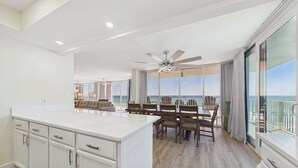 Un vaste espace de vie comprenant un grand îlot central, une table à manger avec des chaises, et un balcon offrant une vue sur l’océan.