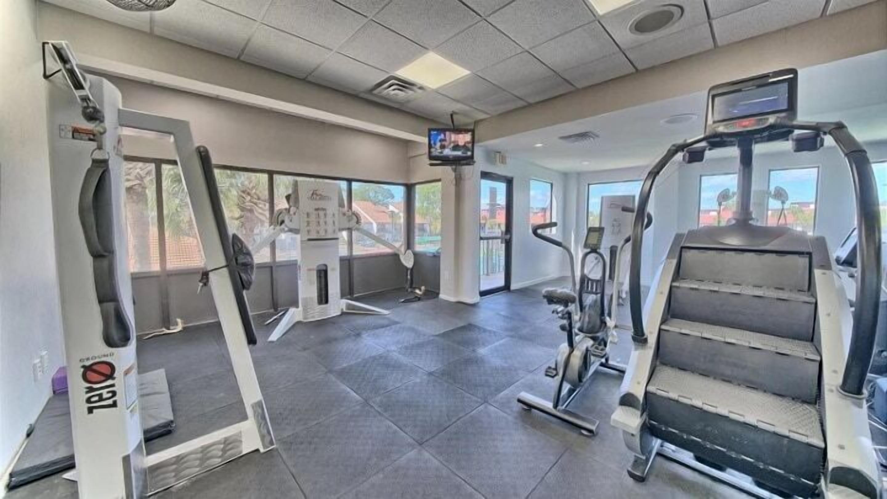 A gym with various exercise equipment, a television screen, and large windows.