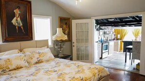 Une chambre à coucher avec un grand lit, une table de chevet, une lampe et une porte donnant sur une cuisine et un coin repas.