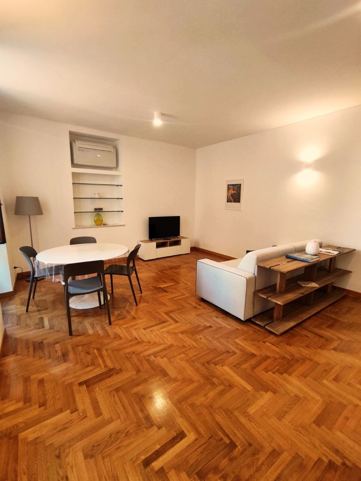 Una sala de estar moderna con un sofá blanco, una mesa de centro de madera, una mesa de comedor con sillas, un televisor de pantalla plana y un aire acondicionado instalado en la pared.