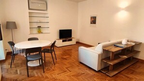 Ein modernes Wohnzimmer mit einem weißen Sofa, einem Holztisch, einem Esstisch mit Stühlen, einem Flachbildfernseher und einer wandmontierten Klimaanlage.