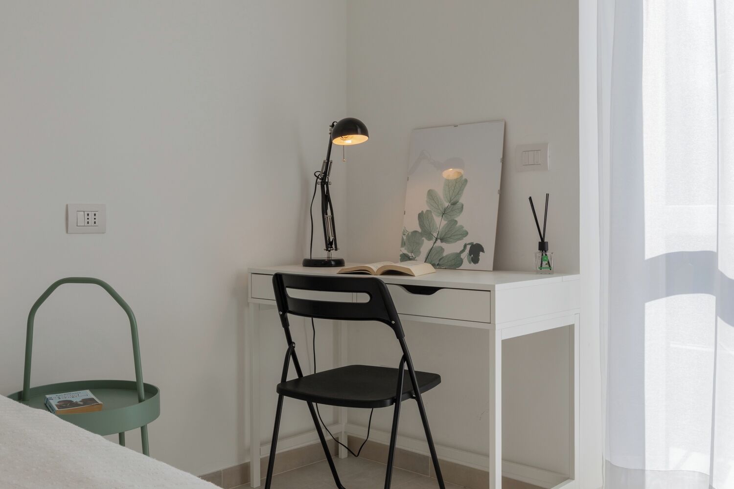 A minimalist bedroom with a white desk, a green chair, a black desk lamp, and a book.