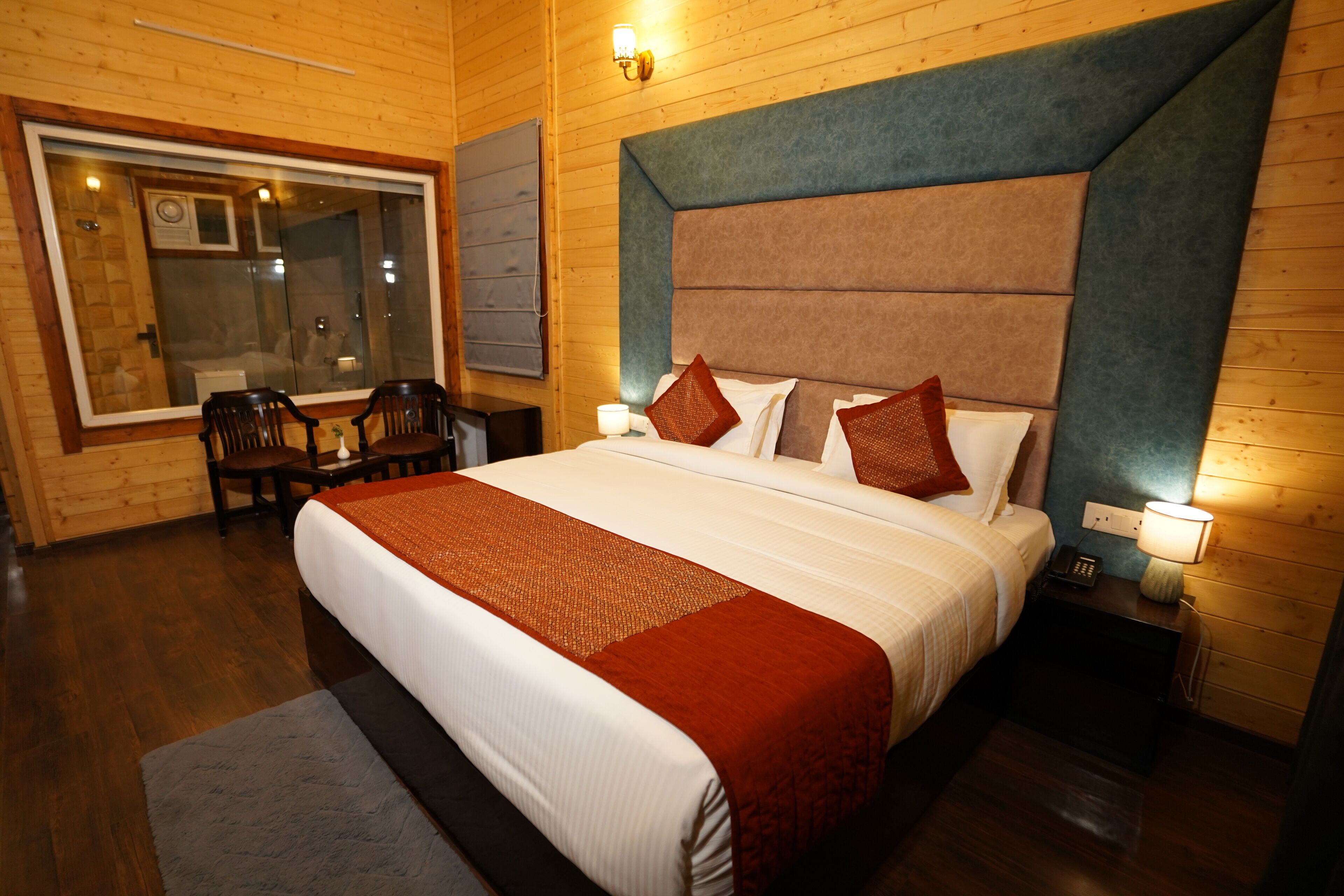 A hotel room with a large bed, wooden walls, a window with blinds, a desk, and a chair.