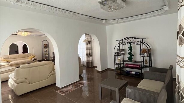 A spacious living area with a large sofa, a smaller sofa, and a coffee table. There are decorative items on shelves and a chandelier on the ceiling. - 4-bedroom Retreat With Lush Gardens Pool- 2346 (Harare)